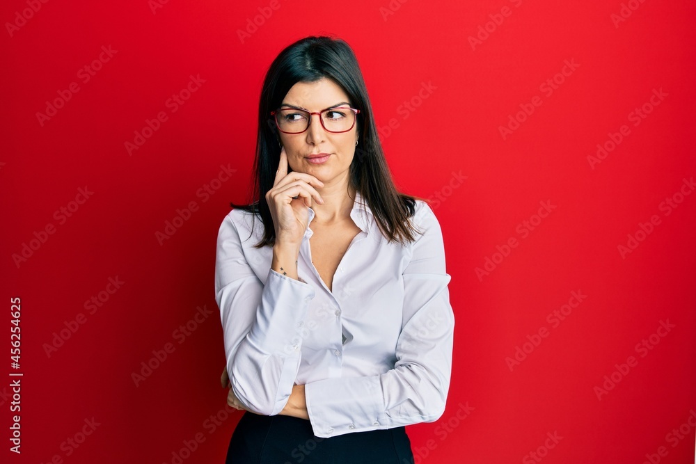 Young hispanic woman wearing business shirt and glasses with hand on chin thinking about question, pensive expression. smiling with thoughtful face. doubt concept. Young hispanic woman wearing business shirt and glasses with hand on chin thinking about question, pensive expression. smiling with thoughtful face. doubt concept.