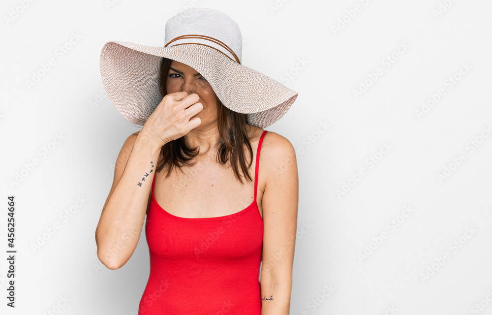 Beautiful brunette woman wearing swimsuit and summer hat smelling something stinky and disgusting, intolerable smell, holding breath with fingers on nose. bad smell Beautiful brunette woman wearing swimsuit and summer hat smelling something stinky and disgusting, intolerable smell, holding breath with fingers on nose. bad smell
