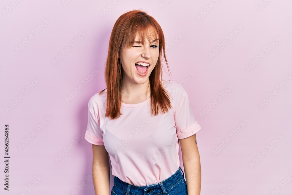 Redhead young woman wearing casual pink t shirt winking looking at the camera with sexy expression, cheerful and happy face. Redhead young woman wearing casual pink t shirt winking looking at the camera with sexy expression, cheerful and happy face.