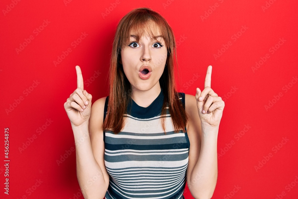 Redhead young woman wearing casual t shirt amazed and surprised looking up and pointing with fingers and raised arms. Redhead young woman wearing casual t shirt amazed and surprised looking up and pointing with fingers and raised arms.