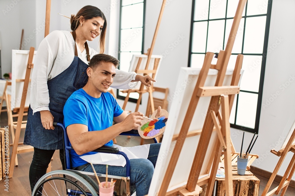 Teacher and disabled paint student sitting on wheelchair smiling happy painting at art school.