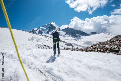 Young female Back view portrait Rope team member on acclimatization day dressed mountaineering clothes walking by snowy slopes in climbing harness and green dynamic rope on the close-up foreground.
