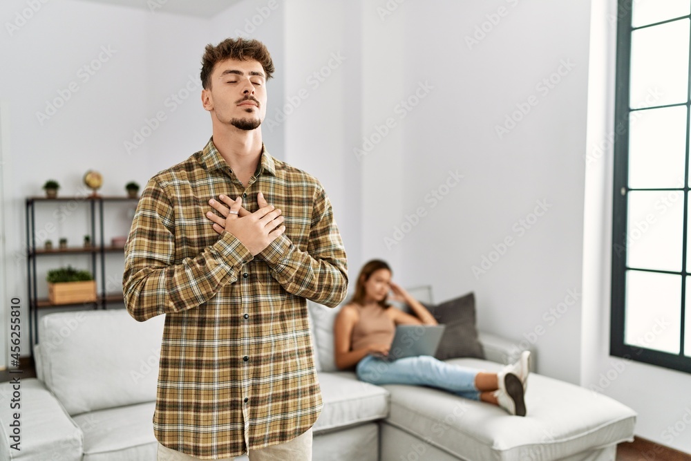 Young handsome man at the living room with girlfriend smiling with hands on chest with closed eyes and grateful gesture on face. health concept. Young handsome man at the living room with girlfriend smiling with hands on chest with closed eyes and grateful gesture on face. health concept.