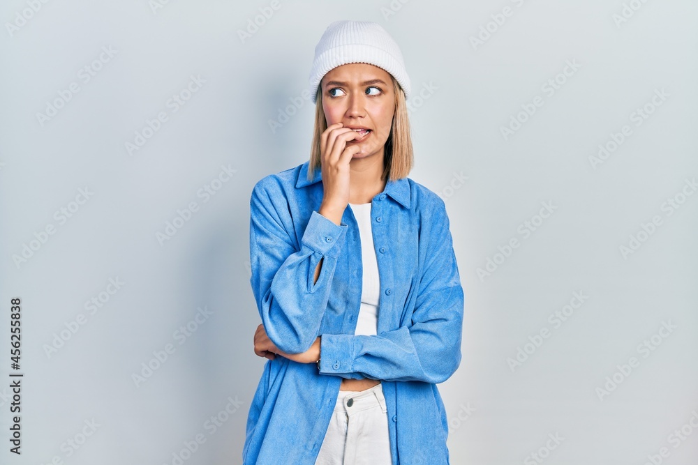 Beautiful blonde woman wearing wool hat looking stressed and nervous with hands on mouth biting nails. anxiety problem. Beautiful blonde woman wearing wool hat looking stressed and nervous with hands on mouth biting nails. anxiety problem.