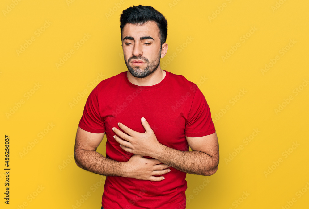 Hispanic man with beard wearing casual red t shirt with hand on stomach because indigestion, painful illness feeling unwell. ache concept. Hispanic man with beard wearing casual red t shirt with hand on stomach because indigestion, painful illness feeling unwell. ache concept.