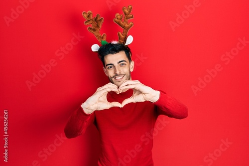 Young hispanic man wearing cute christmas reindeer horns smiling in love doing heart symbol shape with hands. romantic concept.
