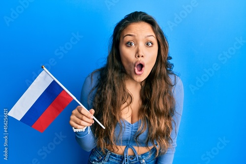 Young hispanic girl holding russia flag scared and amazed with open mouth for surprise  disbelief face