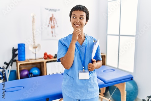 Young hispanic woman with short hair working at pain recovery clinic with hand on chin thinking about question, pensive expression. smiling and thoughtful face. doubt concept.