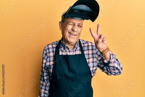 Handsome senior man with grey hair wearing welding protection mask smiling with happy face winking at the camera doing victory sign. number two.
