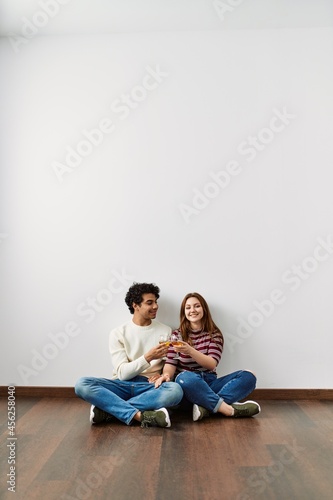 Young hispanic couple toasting with champagne sitting on the floor at empty new home.