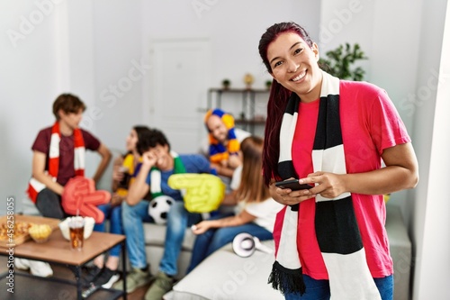 Group of young hispanic friends watching and supporting soccer match. Woman smiling happy and using smartphone at home.