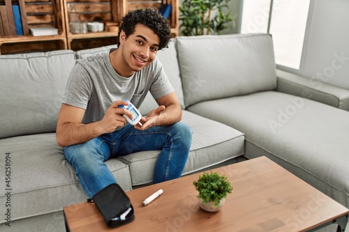 Young hispanic diabetic man measuring glucose sitting on the sofa at home. photo