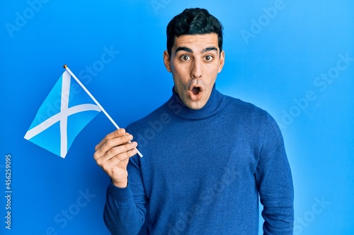 Handsome hispanic man holding scotland flag scared and amazed with open mouth for surprise, disbelief face