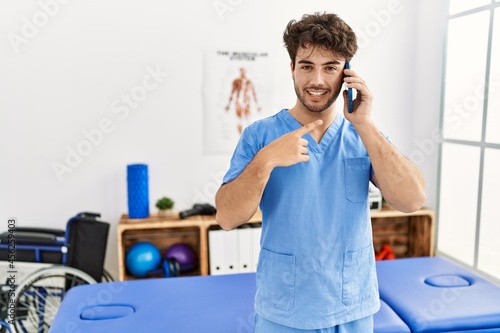 Hispanic physiotherapy man working at pain recovery clinic speaking on the phone smiling happy pointing with hand and finger
