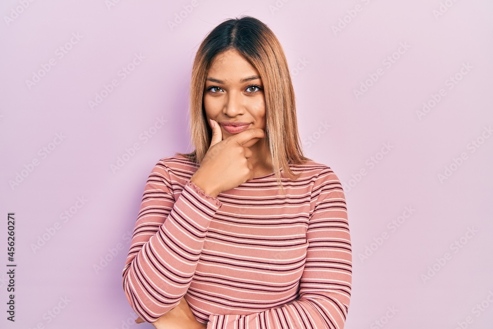 Beautiful hispanic woman wearing casual striped sweater looking confident at the camera smiling with crossed arms and hand raised on chin. thinking positive. Beautiful hispanic woman wearing casual striped sweater looking confident at the camera smiling with crossed arms and hand raised on chin. thinking positive.