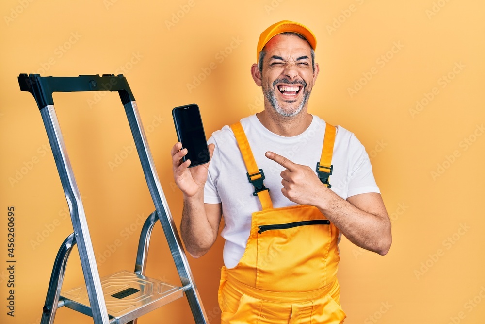 Handsome middle age man with grey hair standing by ladder showing smartphone smiling and laughing hard out loud because funny crazy joke. Handsome middle age man with grey hair standing by ladder showing smartphone smiling and laughing hard out loud because funny crazy joke.