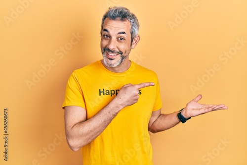 Handsome middle age man with grey hair wearing t shirt with happiness word message amazed and smiling to the camera while presenting with hand and pointing with finger.