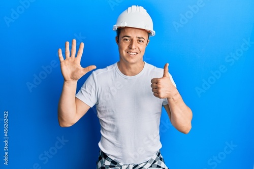 Handsome young man wearing builder uniform and hardhat showing and pointing up with fingers number six while smiling confident and happy.