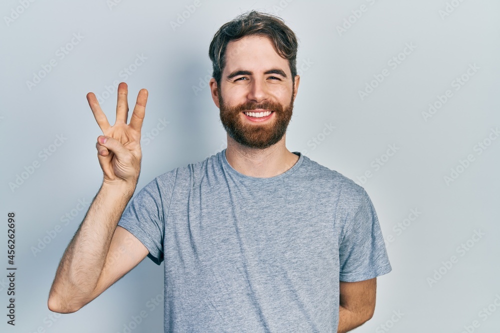 Caucasian man with beard wearing casual grey t shirt showing and pointing up with fingers number three while smiling confident and happy. Caucasian man with beard wearing casual grey t shirt showing and pointing up with fingers number three while smiling confident and happy.