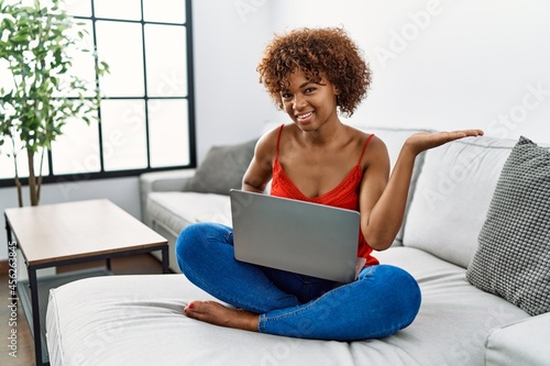 Young african american woman sitting on the sofa at home using laptop smiling cheerful presenting and pointing with palm of hand looking at the camera.