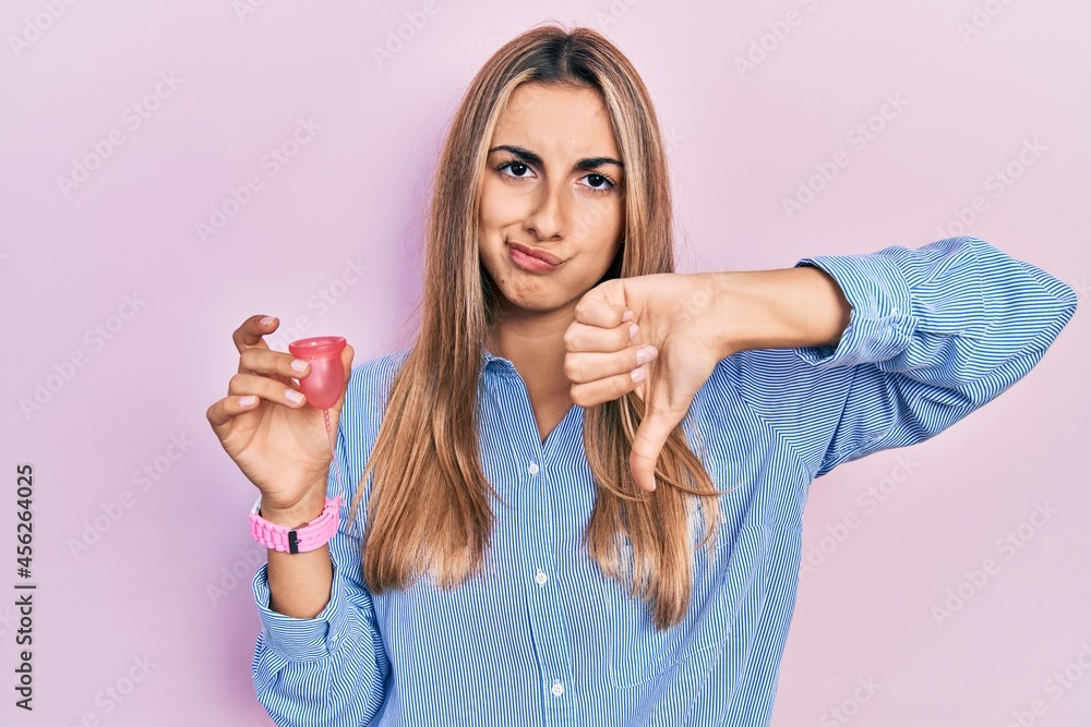 Fototapeta premium Beautiful hispanic woman holding menstrual cup with angry face, negative sign showing dislike with thumbs down, rejection concept