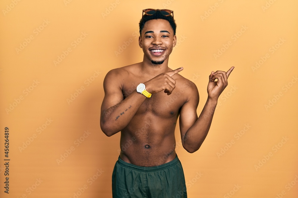 Young african american man wearing swimwear and swimmer glasses smiling and looking at the camera pointing with two hands and fingers to the side.