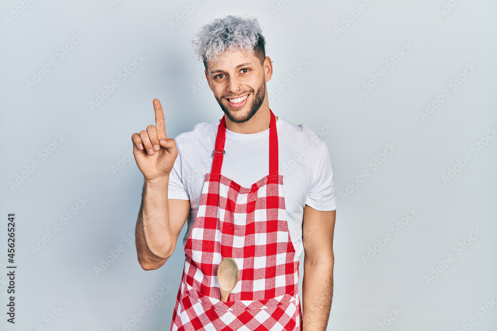 Young hispanic man with modern dyed hair wearing apron showing and pointing up with finger number one while smiling confident and happy. Young hispanic man with modern dyed hair wearing apron showing and pointing up with finger number one while smiling confident and happy.