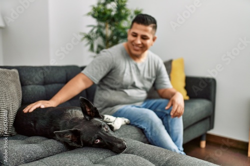 Young hispanic man smiling happy sitting on the sofa with dog at home. © Krakenimages.com