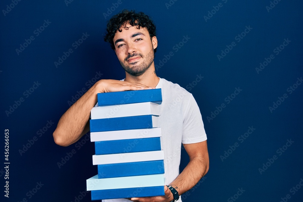 Young hispanic man holding a pile of books relaxed with serious expression on face. simple and natural looking at the camera. Young hispanic man holding a pile of books relaxed with serious expression on face. simple and natural looking at the camera.