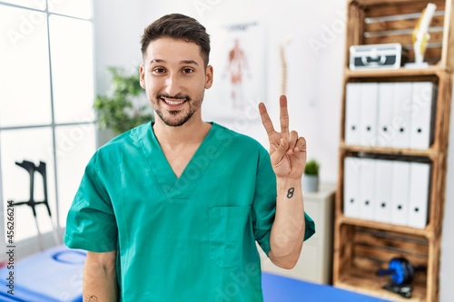 Young physiotherapist man working at pain recovery clinic smiling looking to the camera showing fingers doing victory sign. number two.