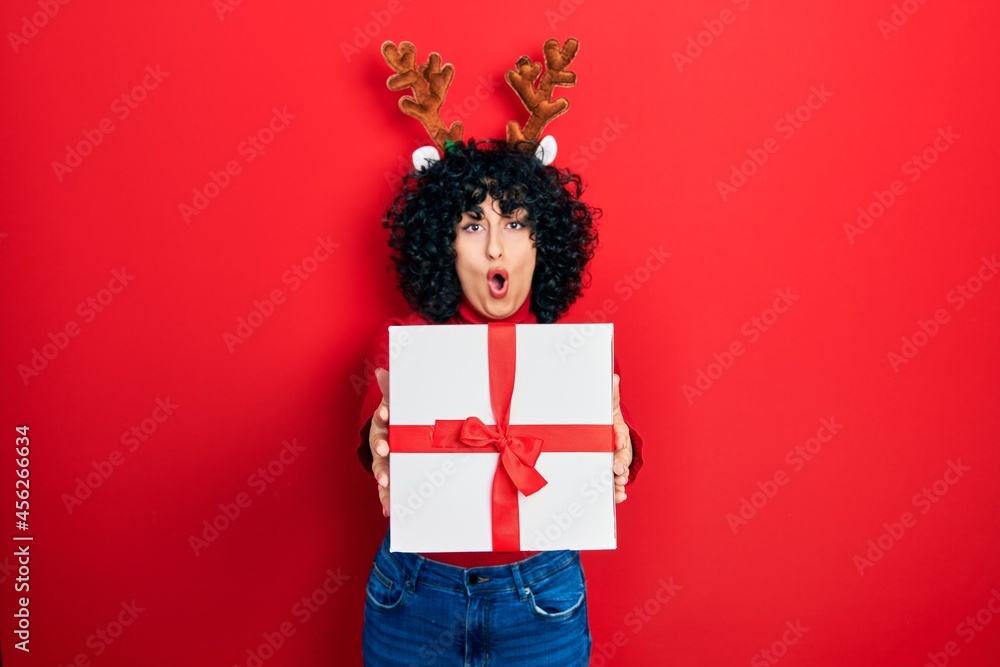 Young middle east woman wearing deer christmas hat holding gift afraid and shocked with surprise and amazed expression, fear and excited face. Young middle east woman wearing deer christmas hat holding gift afraid and shocked with surprise and amazed expression, fear and excited face.