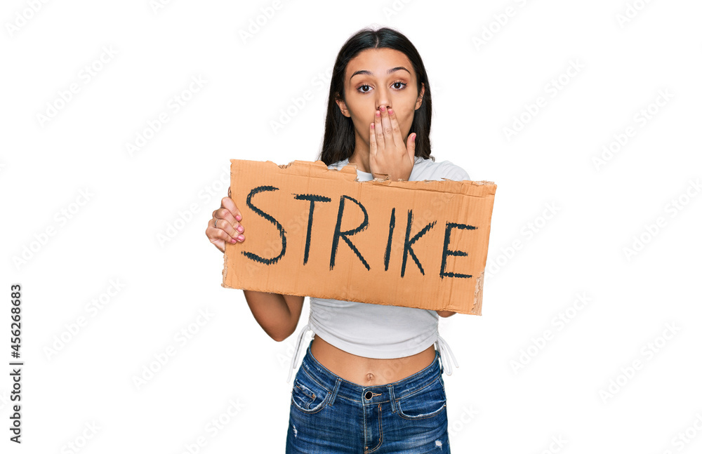 Young hispanic girl holding strike banner cardboard covering mouth with hand, shocked and afraid for mistake. surprised expression Young hispanic girl holding strike banner cardboard covering mouth with hand, shocked and afraid for mistake. surprised expression
