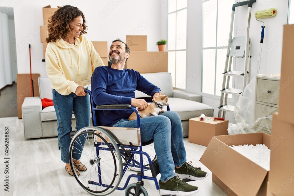 Fototapeta premium Middle age hispanic couple smiling happy. Man sitting on wheelchair with dog on his legs at new home.
