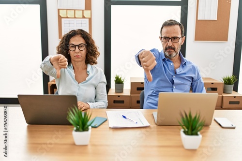 Middle age hispanic woman and man sitting with laptop at the office looking unhappy and angry showing rejection and negative with thumbs down gesture. bad expression.