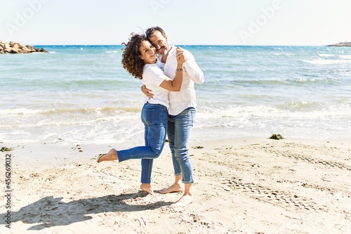 Middle age hispanic couple smiling happy and hugging standing at the beach.