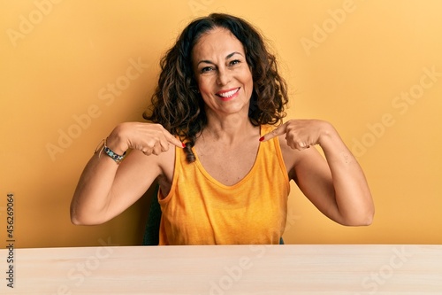 Beautiful middle age woman wearing casual clothes sitting on the table looking confident with smile on face, pointing oneself with fingers proud and happy. © Krakenimages.com