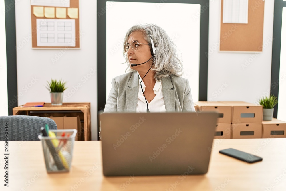 Middle age businesswoman sitting on desk working using laptop at office looking to side, relax profile pose with natural face with confident smile. Middle age businesswoman sitting on desk working using laptop at office looking to side, relax profile pose with natural face with confident smile.