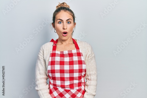 Beautiful caucasian woman with blond hair wearing apron scared and amazed with open mouth for surprise, disbelief face