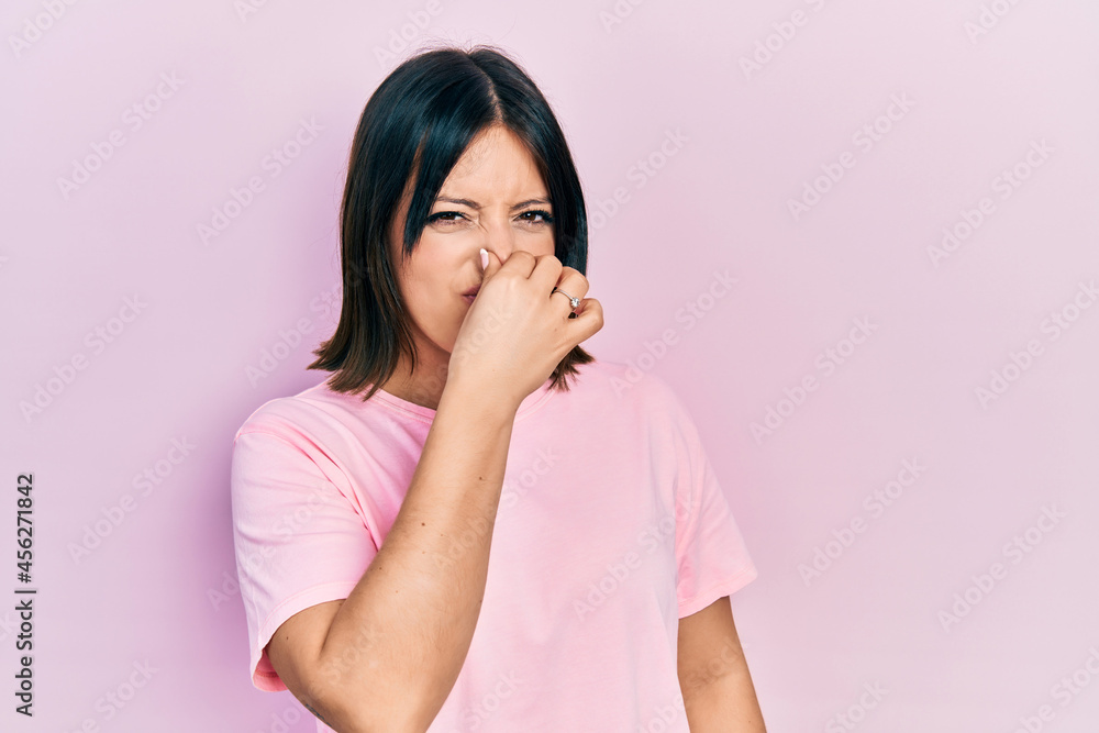 Young hispanic woman wearing casual pink t shirt smelling something stinky and disgusting, intolerable smell, holding breath with fingers on nose. bad smell Young hispanic woman wearing casual pink t shirt smelling something stinky and disgusting, intolerable smell, holding breath with fingers on nose. bad smell
