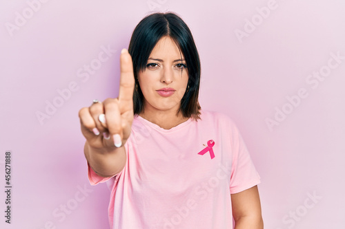 Young hispanic woman wearing pink cancer ribbon on t shirt pointing with finger up and angry expression  showing no gesture