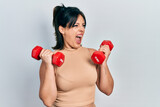 Young hispanic woman wearing sportswear using dumbbells angry and mad screaming frustrated and furious, shouting with anger. rage and aggressive concept.