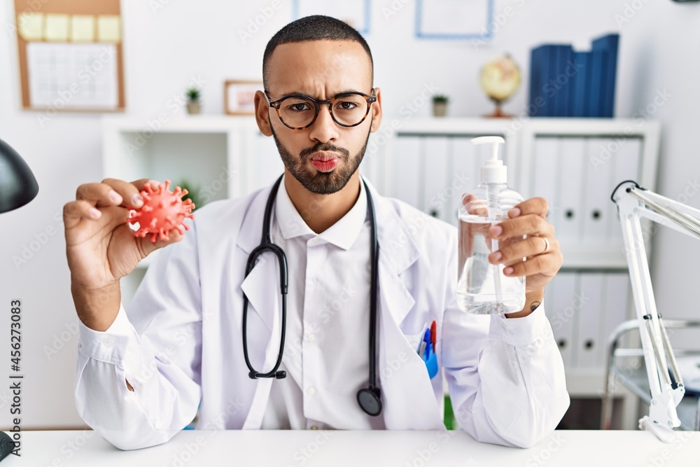 African american doctor man holding virus toy an sanitizer gel puffing cheeks with funny face. mouth inflated with air, catching air. African american doctor man holding virus toy an sanitizer gel puffing cheeks with funny face. mouth inflated with air, catching air.