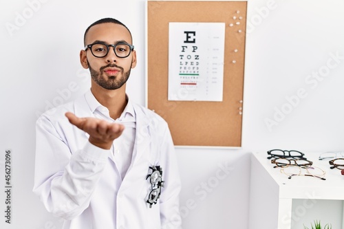 African american optician man standing by eyesight test looking at the camera blowing a kiss with hand on air being lovely and sexy. love expression.