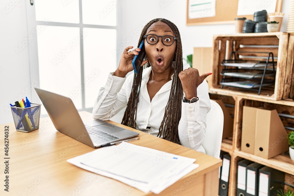 Black woman with braids working at the office speaking on the phone surprised pointing with hand finger to the side, open mouth amazed expression. Black woman with braids working at the office speaking on the phone surprised pointing with hand finger to the side, open mouth amazed expression.
