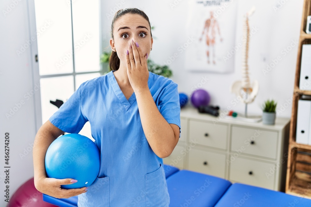 Young physiotherapist woman holding pilates ball at medical clinic covering mouth with hand, shocked and afraid for mistake. surprised expression Young physiotherapist woman holding pilates ball at medical clinic covering mouth with hand, shocked and afraid for mistake. surprised expression