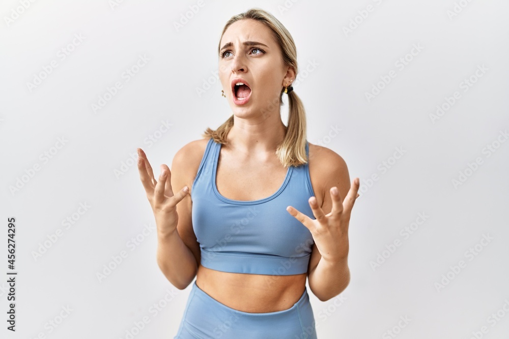 Young blonde woman wearing sportswear over isolated background crazy and mad shouting and yelling with aggressive expression and arms raised. frustration concept. Young blonde woman wearing sportswear over isolated background crazy and mad shouting and yelling with aggressive expression and arms raised. frustration concept.