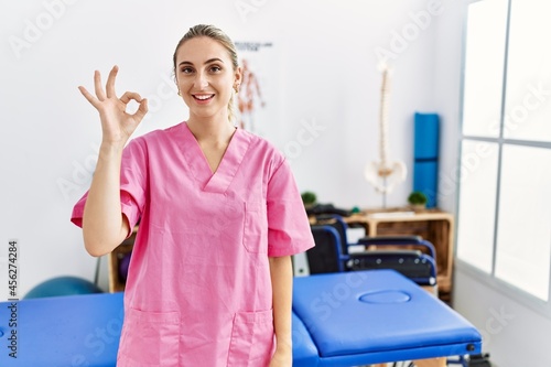 Young blonde woman working at pain recovery clinic smiling positive doing ok sign with hand and fingers. successful expression.