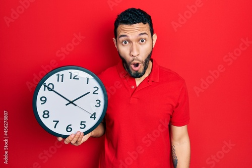 Hispanic man with beard holding big clock scared and amazed with open mouth for surprise, disbelief face