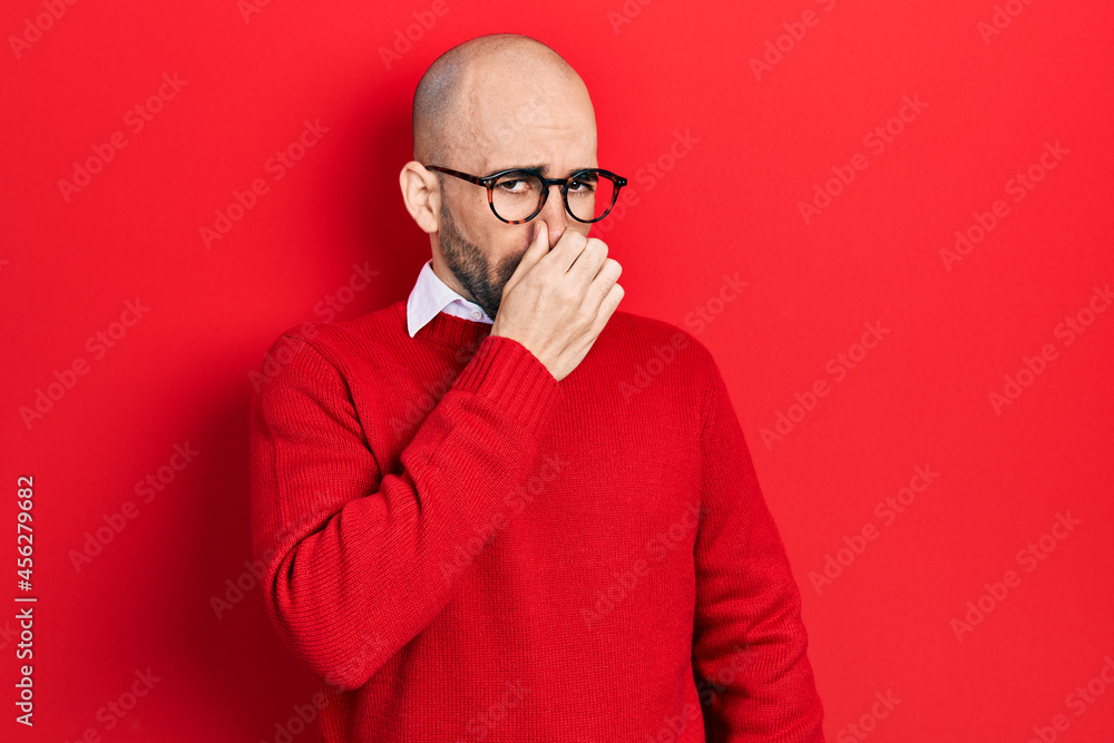 Young bald man wearing casual clothes and glasses smelling something stinky and disgusting, intolerable smell, holding breath with fingers on nose. bad smell Young bald man wearing casual clothes and glasses smelling something stinky and disgusting, intolerable smell, holding breath with fingers on nose. bad smell