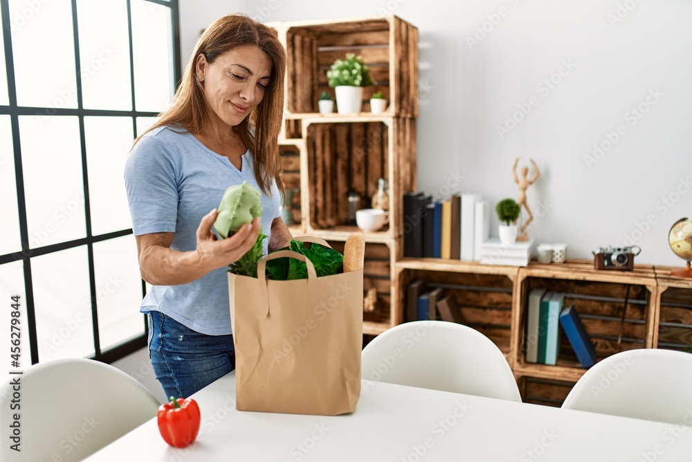 Middle age caucasian woman holding paper bag with groceries standing at home.